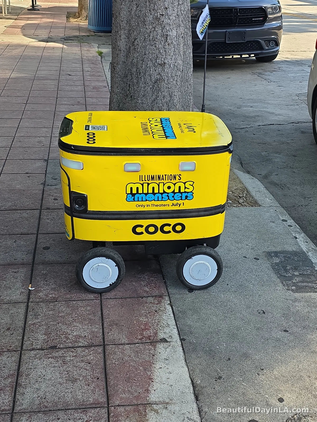 A bright yellow Coco delivery robot parked on a Westwood Village sidewalk